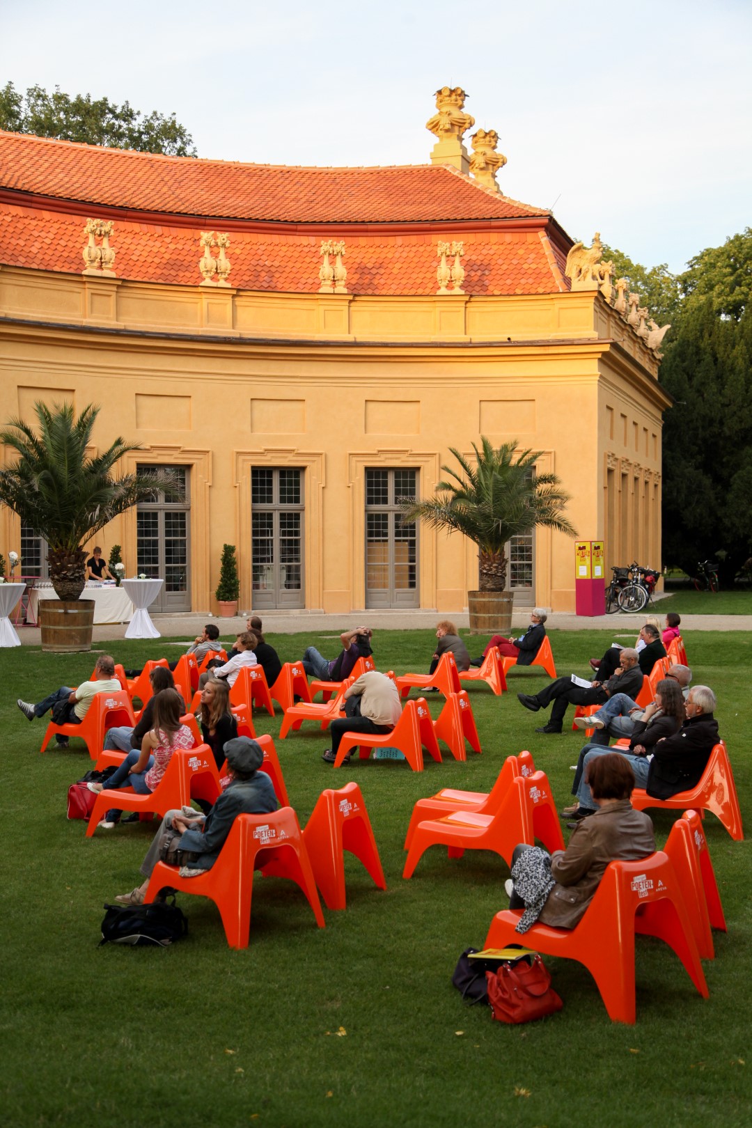 Zuhörer im Schlossgarten sitzen auf roten Liegen. Im Hintergrund die Orangerie