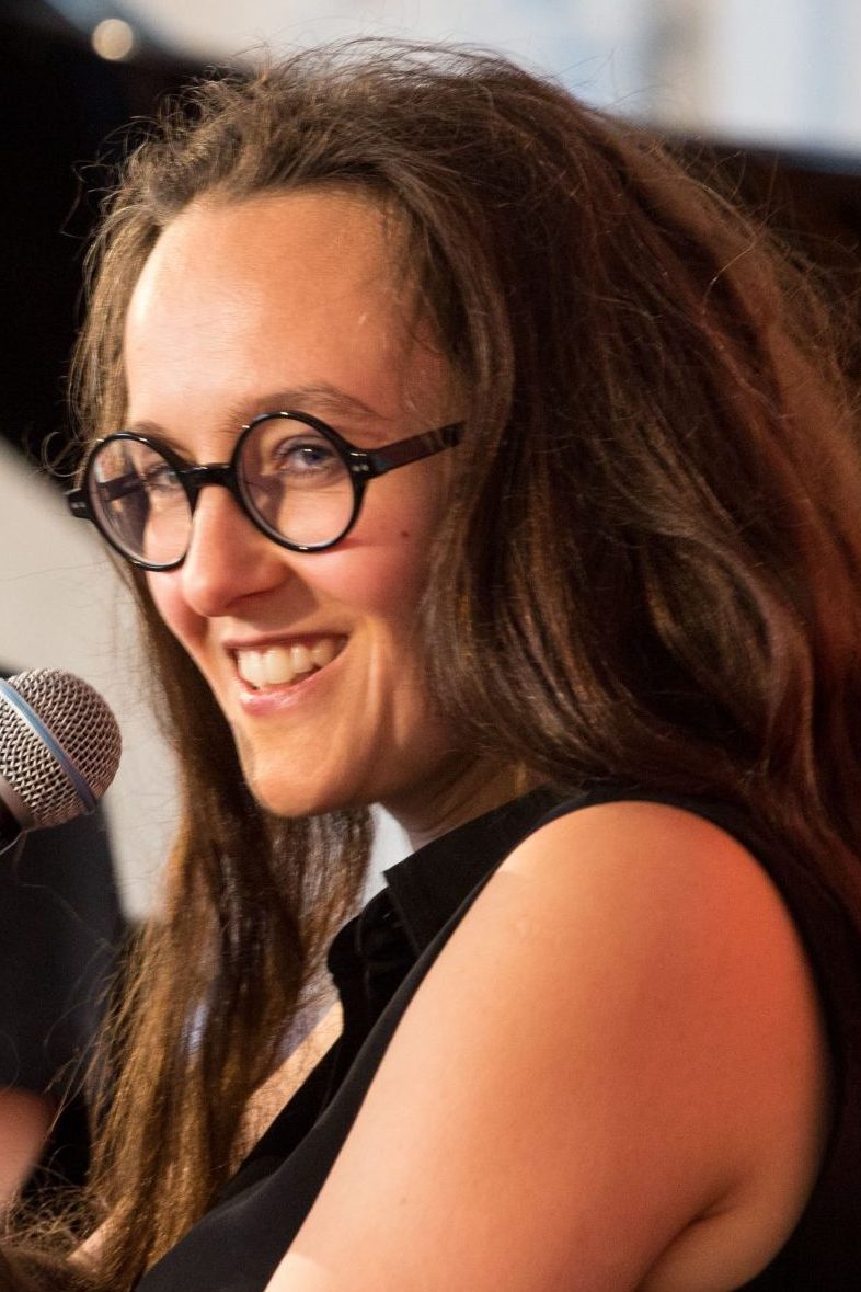 Frau mit Brille und langen, braunen Haaren lächelnd mit Mikrofon in der Hand