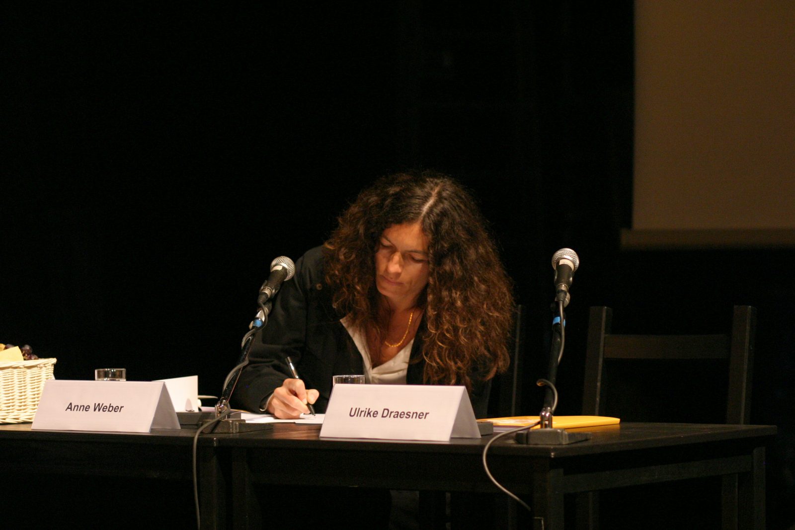 Frau mit langen braunen Haaren sitzt am Podium und schreibt auf ein Blatt Papier