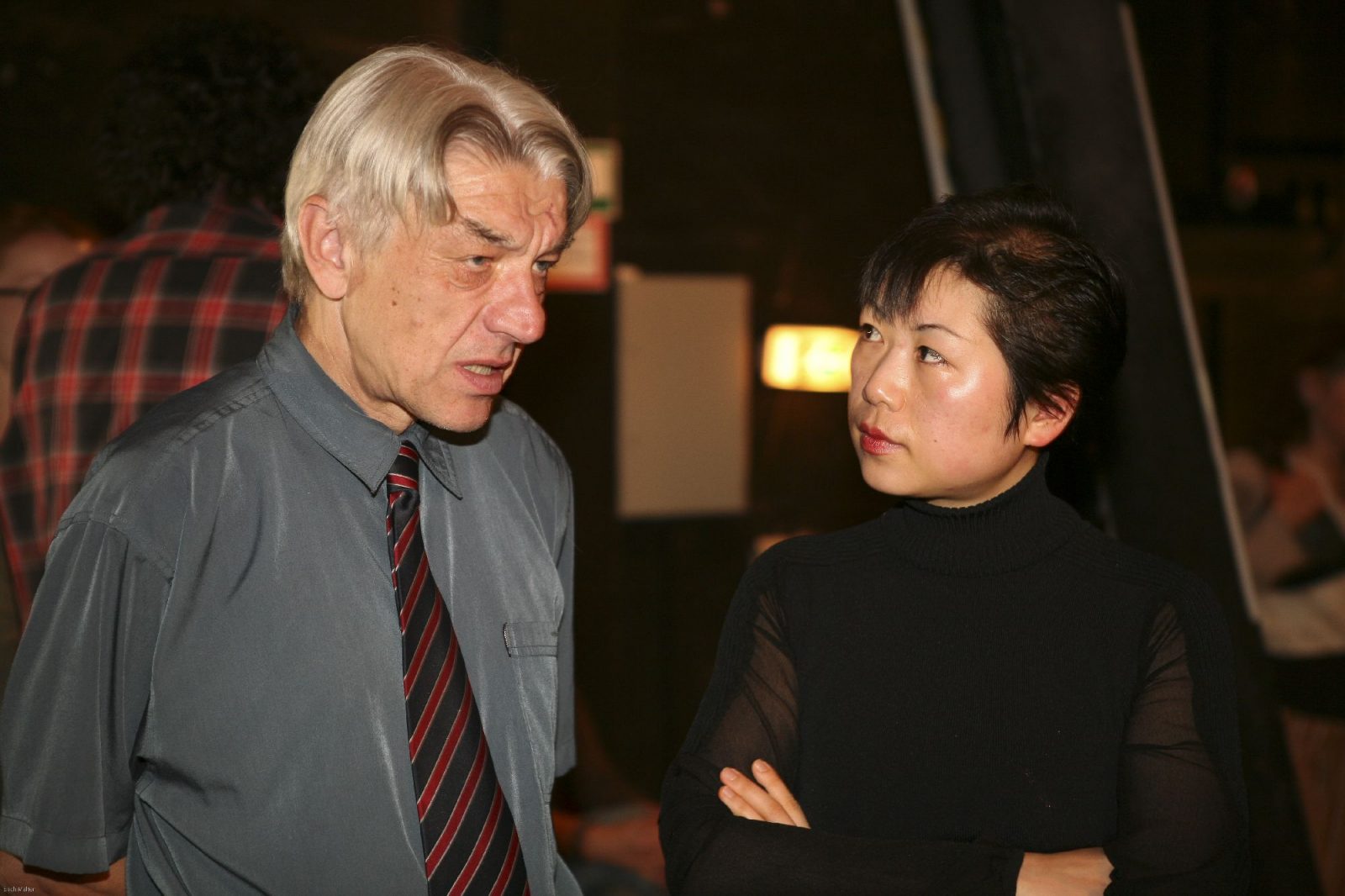 Älterer Mann mit Hemd und Krawatte im Gespräch mit einer asiatiscehn Frau in Schwarz und kurzen Haaren.